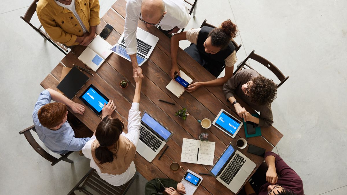 Vista cenital de un equipo reunido alrededor de una mesa de madera con portátiles, tabletas y móviles encendidos mientras dos personas se dan la mano